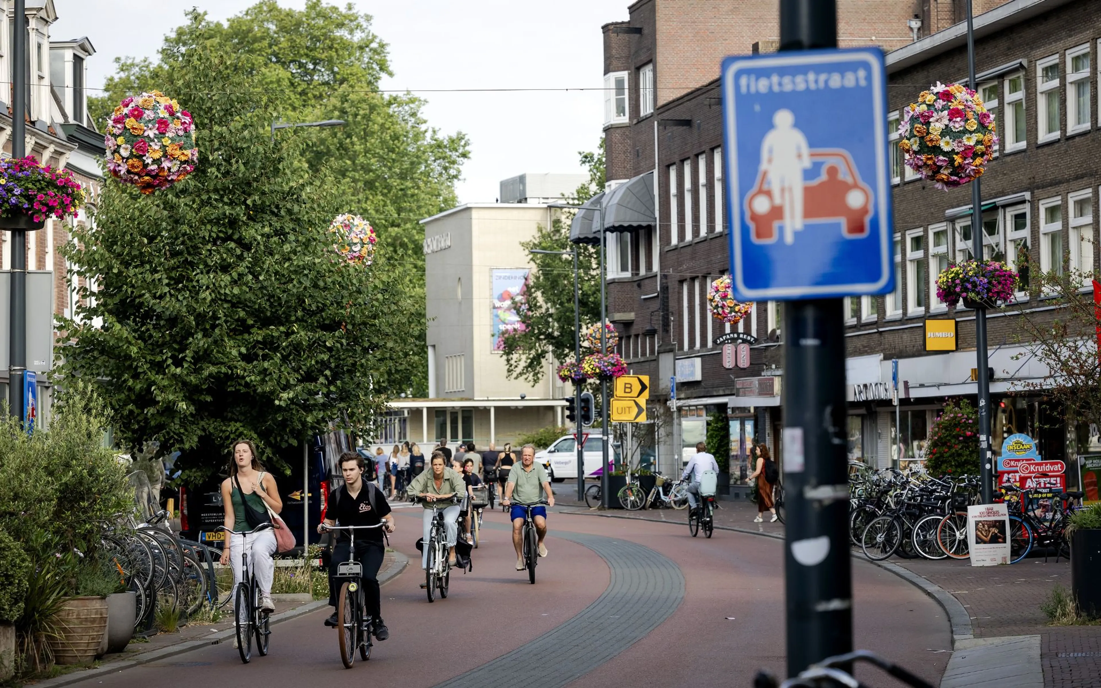 Fietsstraat (Nederlandse winkelstraat als fietsstraat) — een drukke straat ingericht voor fietsverkeer, met een blauw verkeersbord “fietsstraat” en een rood geverfde weg terwijl meerdere fietsers in richting van de camera rijden tussen bakstenen gebouwen en winkelgevels. Langs beide kanten staan veel geparkeerde fietsen en hangen bolvormige bloemmanden vol bloemen, wat bijdraagt aan een zomerse, gezellige uitstraling.