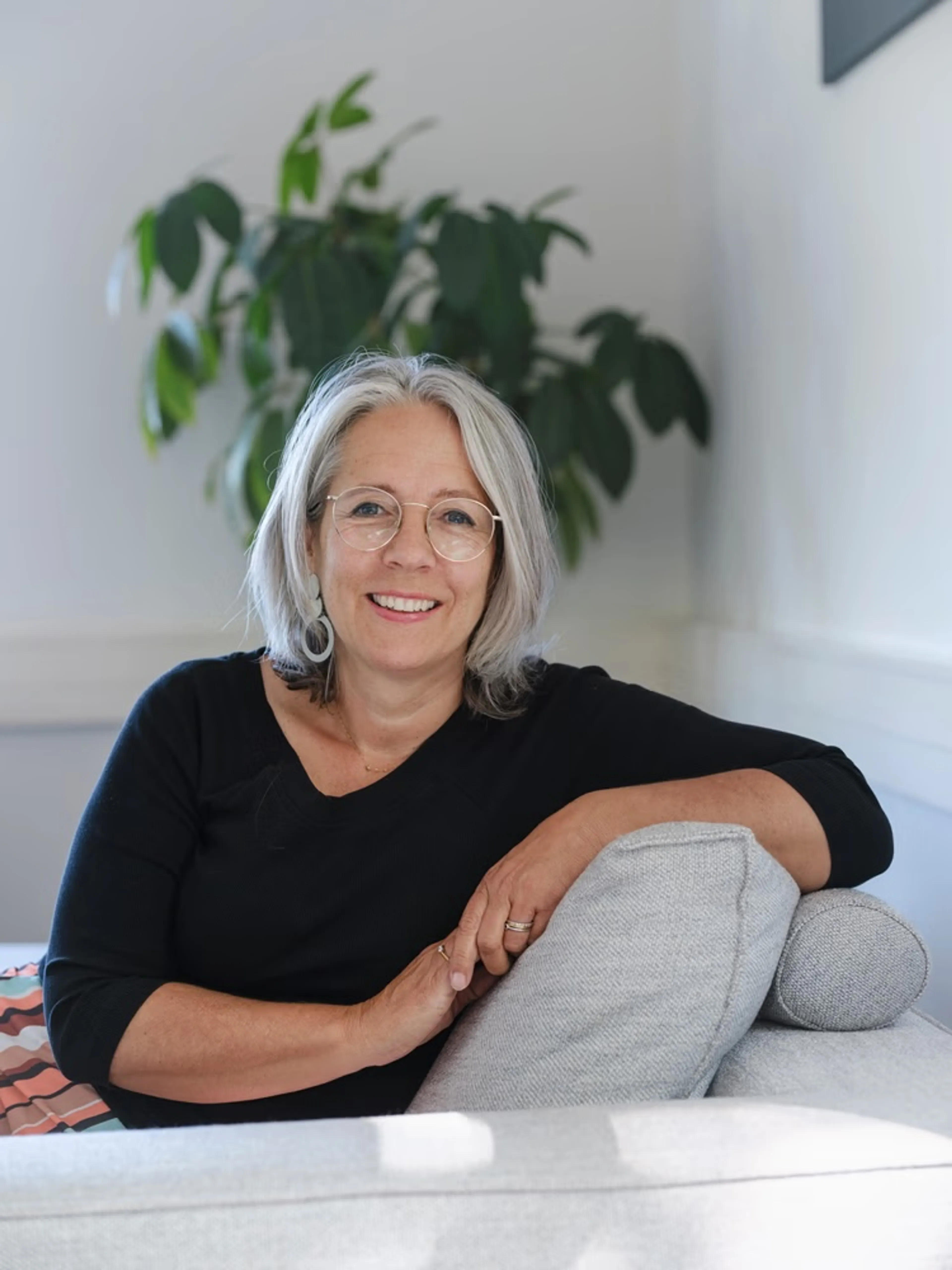 Een middelbare vrouw met zilvergrijs schouderlang haar en ronde bril zit ontspannen op een lichtgrijze bank in een rustige woonkamer; ze draagt een zwarte top met driekwartmouwen en grote lichte oorbellen, en haar gezicht toont een vriendelijke glimlach.