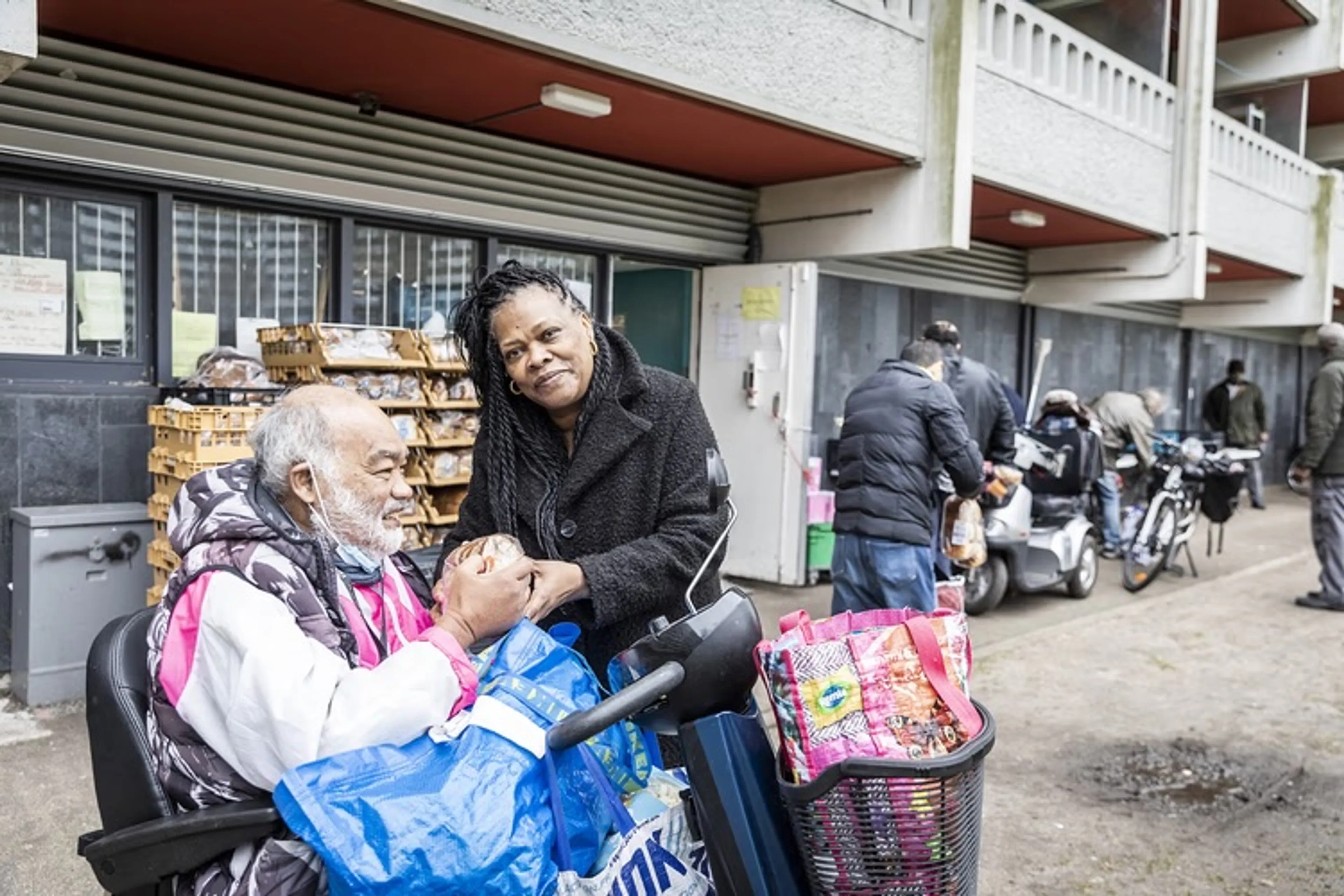 Op een druk uitgiftepunt in een woonwijk is een oudere man op een scootmobiel zichtbaar bezig met eten of vasthouden boven een grote blauwe draagtas, terwijl een middelbare vrouw met vlechten en een dikke zwarte jas hem aandachtig benadert. Om hem heen liggen meerdere tassen en een mand vol kleurrijke boodschappen, met kratten brood en gebak bij ramen met tralies, wat de activiteit en beweging van mensen die spullen inladen benadrukt.