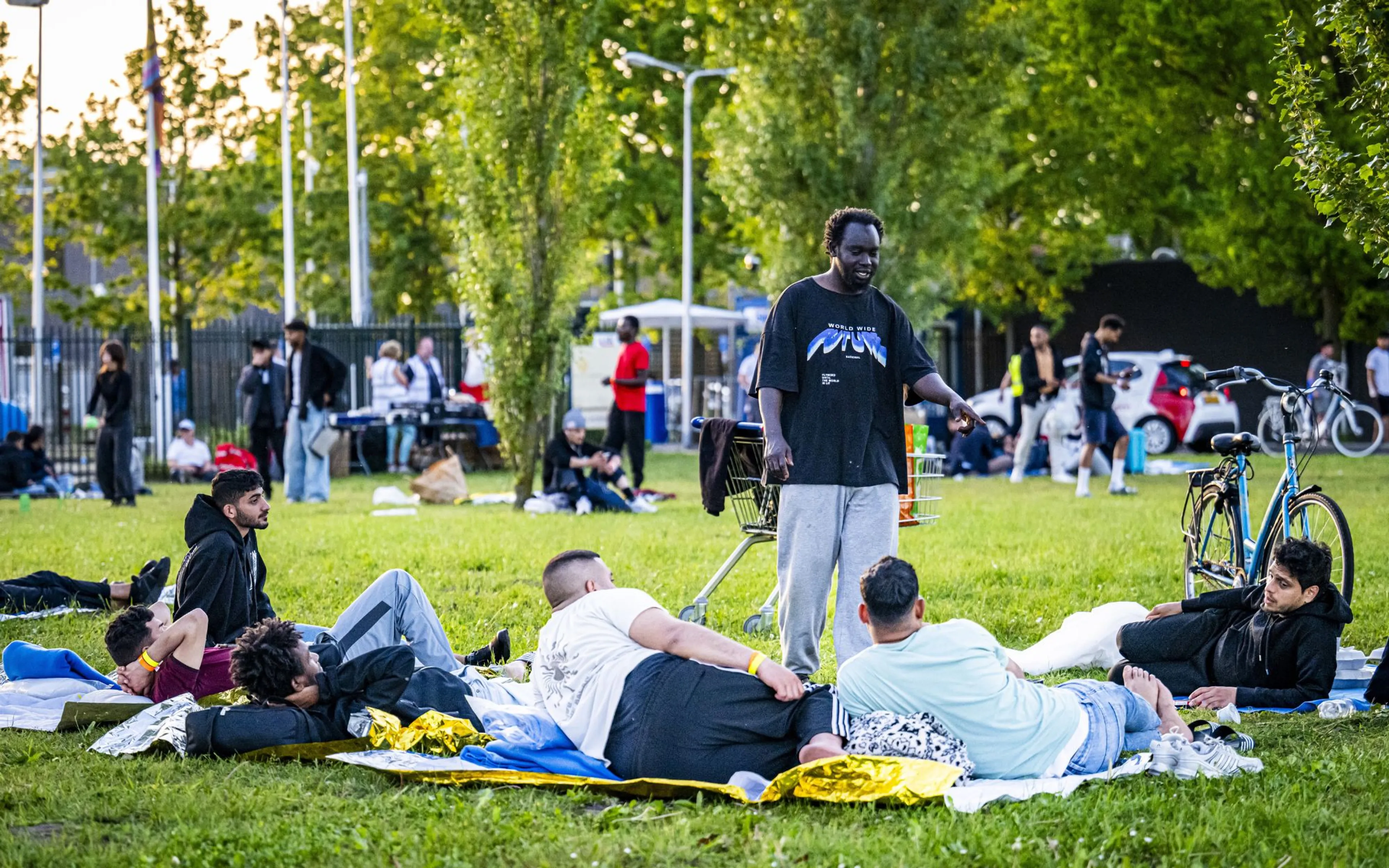 Een groep jongvolwassen mannen ligt en zit op dekens en losse doeken op een grasveld in een groen stadspark, met persoonlijke spullen verspreid om hen heen en één man staand in een zwart T-shirt die lijkt te praten; naast hem staat een winkelwagentje met bagage. In de achtergrond zijn mensen in kleine groepjes, bomen en een hek zichtbaar, met twee fietsen rechts en een kleine auto verderop.