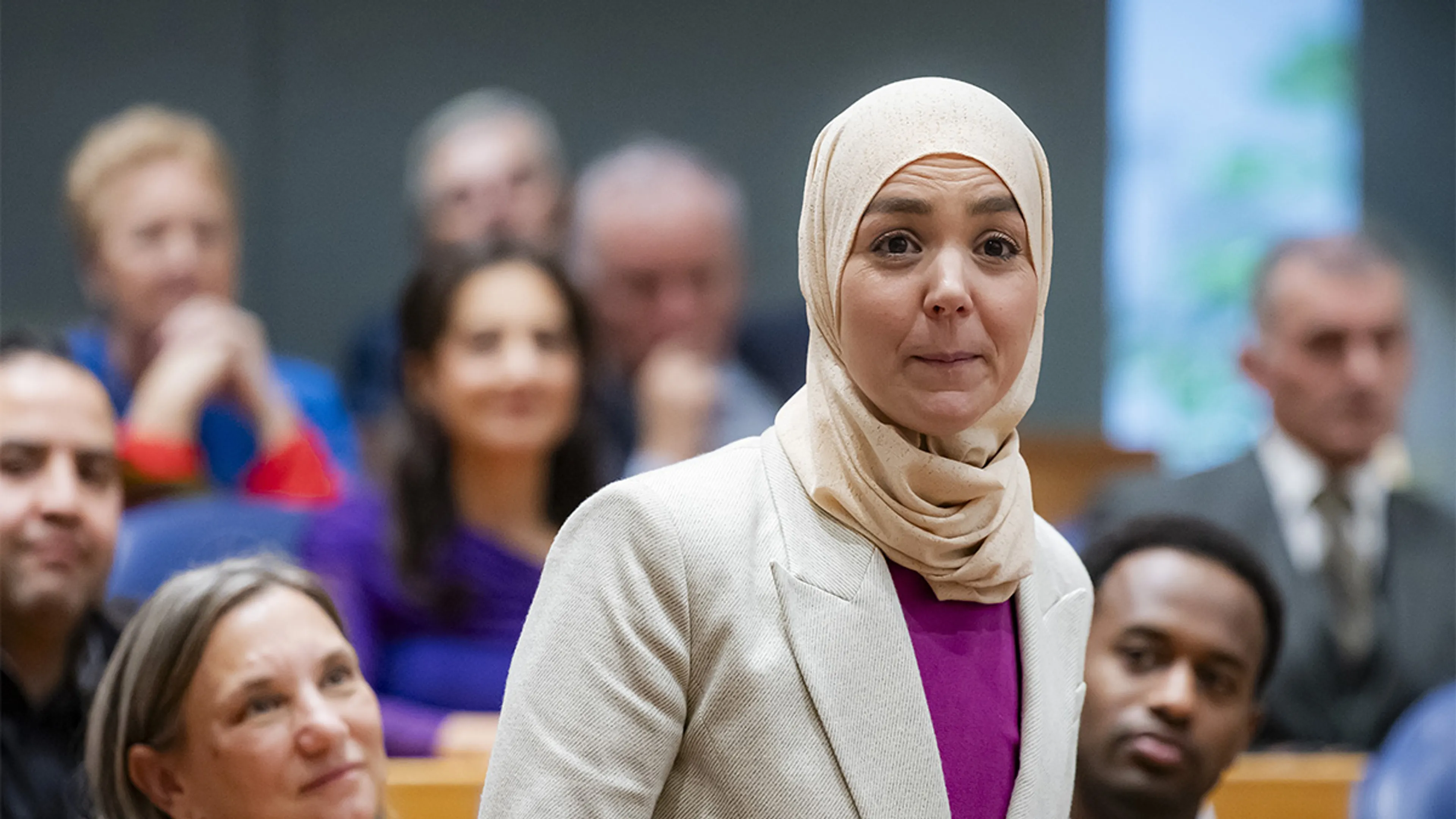 Raadszaalmoment: Een vrouw staat op de voorgrond in een formele vergaderzaal en spreekt, met een serieuze, geconcentreerde uitdrukking; de setting suggereert een officiële discussie terwijl meerdere aanwezigen op houten zitplaatsen luisteren.